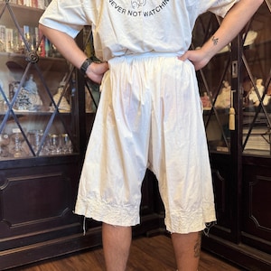 Puede incluir: Bombachos color crema hasta la rodilla con dobladillo bordado. Los bombachos están hechos de una tela ligera y tienen un ajuste holgado y cómodo. La cintura es elástica. La persona lleva una camiseta blanca con el texto "NEVER NOT WATCHING".