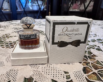 May include: A vintage perfume bottle with a silver cap and a white label that reads "Quadrille Balenciaga Paris France". The bottle is sitting on a white box with a white label that reads "Quadrille Balenciaga Paris".