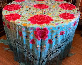 May include: A round, light blue tablecloth featuring vibrant, large embroidered red peony flowers and smaller multi-colored floral accents. The edges of the fabric are finished with a long, matching light blue knotted fringe trim.