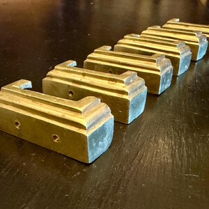 Peut inclure: Un ensemble de six poignées de tiroir rectangulaires en laiton disposées en diagonale. Chaque poignée présente un bord supérieur étagé et deux trous de vis circulaires sur la face avant. Le métal a une finition dorée polie avec une patine visible.