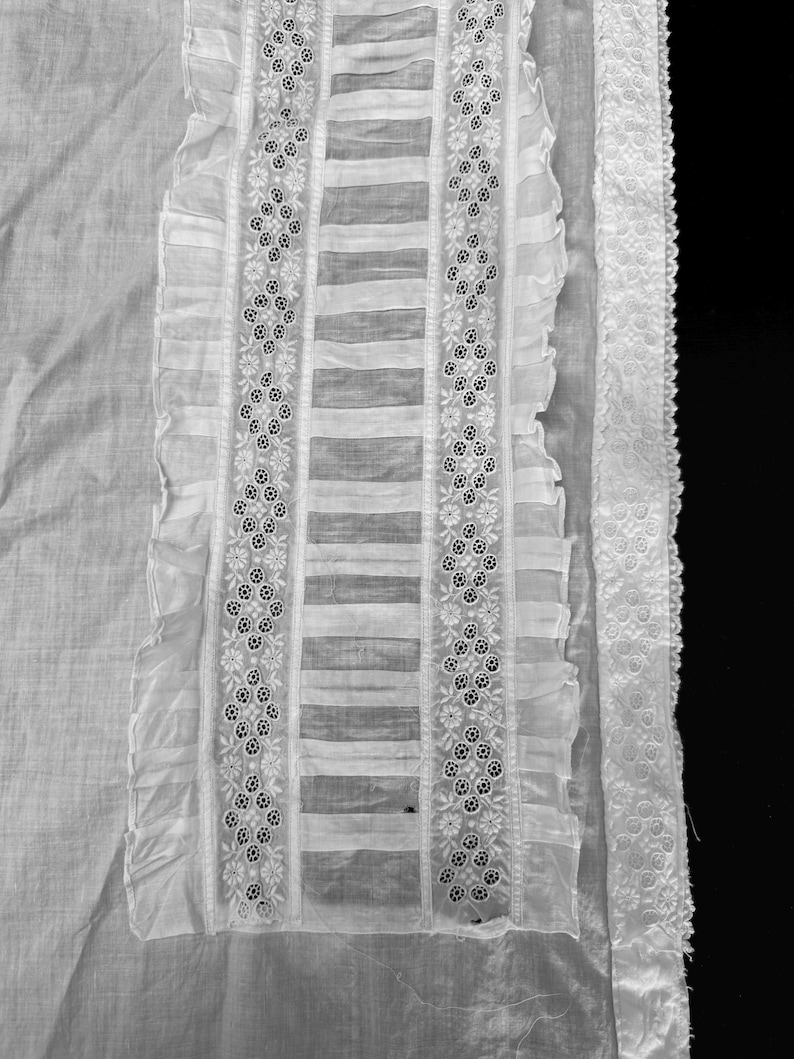May include: White cotton fabric with a delicate lace trim and embroidered details. The fabric has a sheer, almost translucent quality, and the lace is intricate and delicate. The embroidery is in a floral pattern, and the overall effect is very feminine and elegant.