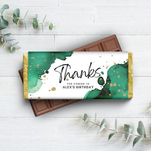 Puede incluir: Una barra de chocolate envuelta en un papel blanco con un diseño de acuarela verde y dorado. El envoltorio dice "Gracias por venir al cumpleaños de Alex".