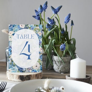 Op de afbeelding: Een blauwe en witte bloemen tafelnummerkaart met de tekst "TABLE 4" op een witte achtergrond. De kaart ligt op een houten schijf.