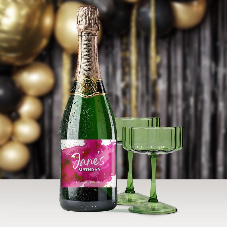 May include: A green glass bottle of sparkling wine with a pink and gold watercolor label that says "Jane's Birthday September 8th 20XX". The bottle is sitting on a white surface next to two green glass champagne flutes.