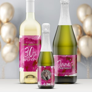 May include: Three bottles of wine with personalized labels for a 30th birthday party. The labels feature a pink watercolor background with gold accents and the text "Welcome to Jane Martin's 30th birthday". One bottle has a photo of a woman on the label.