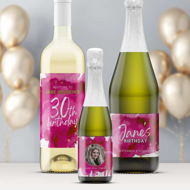 May include: Three bottles of wine with personalized labels for a 30th birthday party. The labels feature a pink watercolor background with gold accents and the text "Welcome to Jane Martin's 30th birthday". One bottle has a photo of a woman on the label.