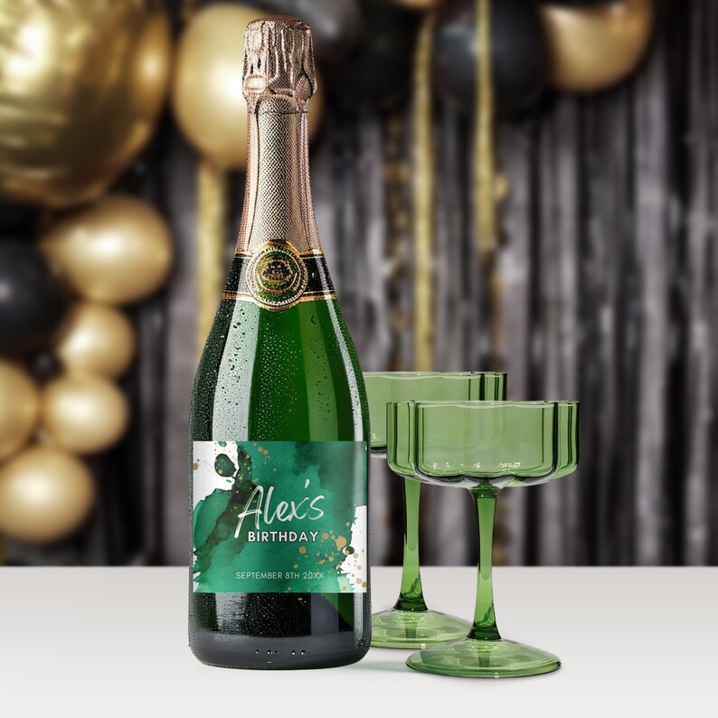 Op de afbeelding: Een groene en gouden champagnefles met een gepersonaliseerd etiket waarop "Alex's Birthday September 8th 20XX" staat. De fles staat naast twee groene champagneflutes.