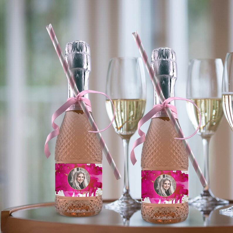 May include: Two small bottles of pink champagne with personalized labels. The labels feature a watercolor design with a photo of a person and the text "Thanks for coming". Each bottle has a pink and white striped straw and a pink ribbon.
