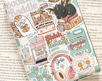 May include: A collection of colorful stickers featuring various bookish themes. The stickers include a ghost reading a book, a dragon sleeping on a stack of books, a laptop with the text "Born to read, forced to work", a pair of headphones with the text "Girlie", a balloon dog with smiley faces, and a bookish fuel sticker with the text "Shhh I'm reading audiobook".