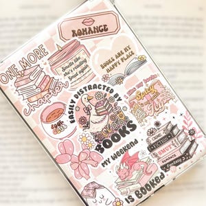 May include: A clear phone case decorated with pink and white stickers. The stickers feature book-related themes, including stacks of books, a candle, a duck, and the words "Romance," "Books," and "Smut." The case has a pink checkered background.