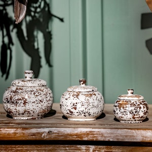 Könnte beinhalten: Drei weiße Keramik-Deckelgläser mit einem abgenutzten Finish. Die Gläser sind in verschiedenen Größen und in einer Reihe auf einer Holzoberfläche angeordnet.