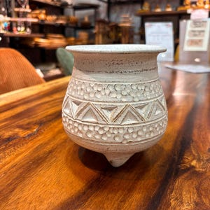Peut inclure: Un petit pot en céramique blanc cassé avec un bord évasé et trois pieds courts. L'extérieur présente une surface texturée avec une bande d'indentations circulaires sculptées au-dessus et au-dessous d'une rangée centrale de triangles incisés.