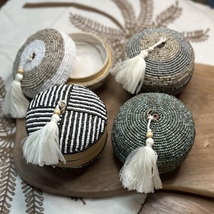 Könnte beinhalten: Vier runde Perlenboxen mit Quasten. Die Boxen sind in Schwarzweiß, Beige und Weiß sowie Blau und Weiß. Die Boxen stehen auf einer Holzoberfläche.