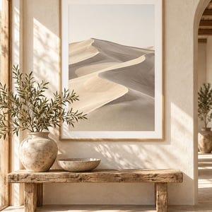 May include: A framed wall art print featuring a desert landscape with rolling sand dunes in shades of beige and soft gray. The artwork is displayed in a light wood frame, hanging on a textured off-white wall above a rustic wooden bench.