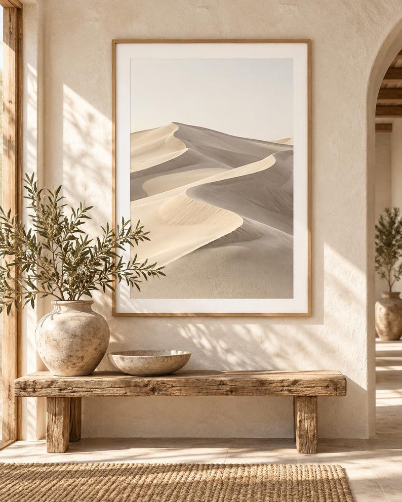 May include: A framed wall art print featuring a desert landscape with rolling sand dunes in shades of beige and soft gray. The artwork is displayed in a light wood frame, hanging on a textured off-white wall above a rustic wooden bench.