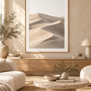 May include: A framed wall art print featuring a desert landscape with rolling sand dunes in shades of beige, tan, and soft gray. The image displays a high-contrast view of sunlit ridges and deep shadows across the sandy terrain.