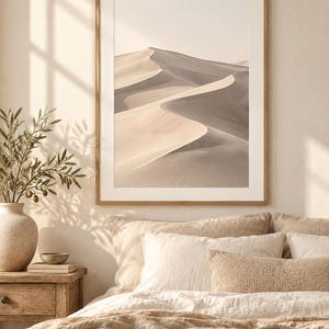May include: A framed wall art print featuring a desert landscape with rolling sand dunes in shades of beige, tan, and soft gray. The image displays a high-contrast view of curved sand ridges under a bright, clear sky, enclosed in a light wood frame.