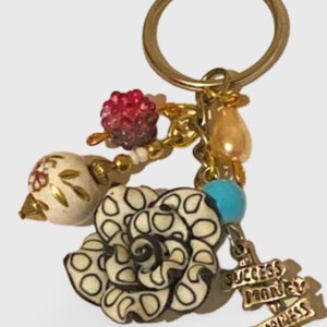 May include: A gold-colored keychain with various charms. Charms include a white and black flower, a red beaded cluster, a teardrop-shaped bead, a blue bead, and a charm with the words "Success, Money, Happiness."