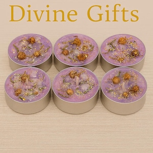 Op de afbeelding: Zes ronde zilverkleurige waxinelichtjes met lavendelkleurige was. Elke kaars is versierd met gedroogde bloemen, gouden glitter en een kleine witte lont. De tekst "Divine Gifts" staat in goud boven de kaarsen.