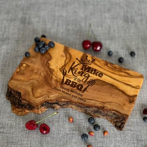 Puede incluir: Una tabla de cortar de madera con borde vivo, grabada con "King of the BBQ" y una corona. La tabla est&aacute; decorada con ar&aacute;ndanos y cerezas. Tambi&eacute;n est&aacute; grabado el texto "Happy Birthday From Mason and Olivia".