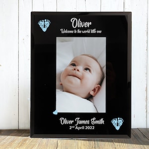 May include: Black glass photo frame with a baby photo inside. The frame has blue footprints and the text "Oliver, Welcome to the world little one, Oliver James Smith, 2nd April 2022".