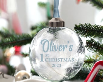 Può includere: Un addobbo natalizio in vetro trasparente con piume bianche all'interno. L'addobbo ha il testo "Oliver's 1st Christmas 2023" scritto in lettere blu.