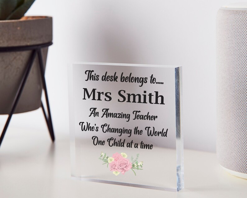 Pu&ograve; includere: Un cartello da scrivania in acrilico trasparente con il testo "This desk belongs to... Mrs Smith An Amazing Teacher Who's Changing the World One Child at a time" e un motivo floreale con fiori rosa.