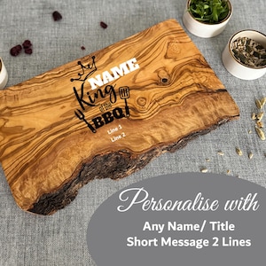 Puede incluir: Una tabla de cortar de madera con borde natural, con el texto "King of the BBQ" y espacio para personalizaci&oacute;n. La tabla es de color marr&oacute;n claro con bordes marrones oscuros y veta de madera natural. Incluye una corona y un gr&aacute;fico de herramientas de barbacoa.