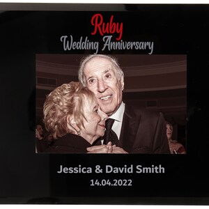 May include: Black framed photo with a picture of an older couple. The text "Ruby Wedding Anniversary" is at the top, with the names "Jessica & David Smith" and the date "14.04.2022" at the bottom.