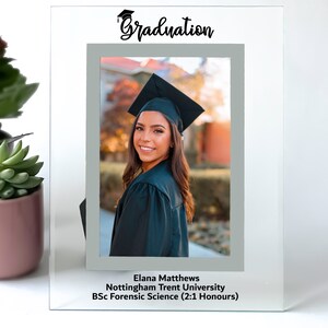 May include: A glass photo frame with a grey mount and a graduation cap icon. The frame has a photo of a woman in a graduation gown and cap. The text "Graduation" is at the top of the frame. The text below the photo reads "Elana Matthews, Nottingham Trent University, BSc Forensic Science (2:1 Honours)"