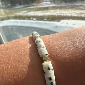 May include: A beaded bracelet featuring white and black speckled disc beads, interspersed with gold-colored square and triangular beads. The bracelet is shown on a person's arm, with a blurred winter landscape in the background.
