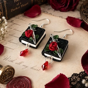 May include: A pair of dangle earrings featuring miniature black books with white pages. Each book is decorated with a small red rose, green leaves, and a silver dagger charm. A faceted red crystal drop hangs from the bottom of each book.