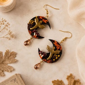 Può includere: Un paio di orecchini a forma di mezzaluna con una silhouette di uccello color oro, piccoli funghi rossi e bianchi, foglie arancioni e bacche gialle su base nera, completati da un cristallo a goccia marrone chiaro e ganci dorati.