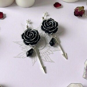 May include: A pair of silver-toned earrings featuring a black rose stud, a dangling black faceted bead, and a silver arrow charm. The earrings are designed with a post back and showcase detailed metalwork on the rose petals and arrow shaft.