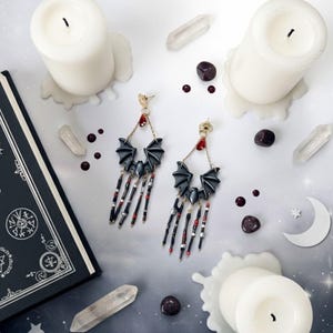 Pendientes hechos a mano con diseño de murciélago en tonos negro y gris, decorados con cuentas colgantes rojas y plateadas. Fondo gris claro con velas blancas, cuarzos, un libro negro y detalles decorativos en forma de luna.