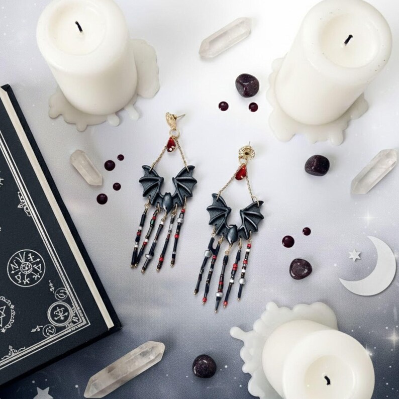 Pendientes hechos a mano con diseño de murciélago en tonos negro y gris, decorados con cuentas colgantes rojas y plateadas. Fondo gris claro con velas blancas, cuarzos, un libro negro y detalles decorativos en forma de luna.