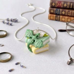Peut inclure: Un collier argenté avec un pendentif représentant un dragon vert sur une pile de livres miniatures. Les livres sont vert pastel et jaune, avec des fleurs blanches. Le fond comprend des livres anciens, des lunettes et des loupes.