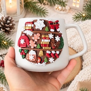 Op de afbeelding: Een witte keramische mok met een cirkelvormige, verhoogde kleidecoratie aan de voorzijde. Het ontwerp toont een bruine houten plank gevuld met miniatuur kerstitems, waaronder peperkoekfiguurtjes, boeken, een rode trui, kousen en een rendier.