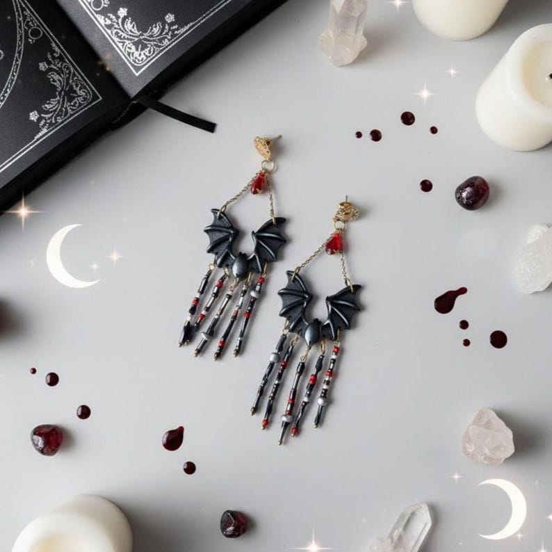 Pendientes hechos a mano con diseño de murciélago en tonos negro y gris, decorados con cuentas colgantes rojas y plateadas. Fondo gris claro con velas blancas, cuarzos, un libro negro y detalles decorativos en forma de luna.