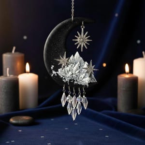 Op de afbeelding: Een hemels thema hangende decoratie met een zwarte halve maan met zilveren ster accenten en een bergketen. Heldere kristalachtige kralen hangen eronder. De achtergrond bevat brandende kaarsen en een donkerblauwe stof.