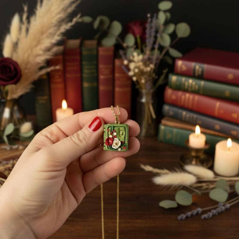 Tres collares con dijes en forma de libro miniatura de arcilla polimérica en colores crema, verde bosque y rojo cereza. Cada libro tiene flores en relieve (rosas y hojas) y una letra dorada en la portada. Las cadenas son de eslabones dorados y están dispuestas en diagonal sobre un fondo liso.