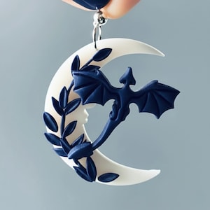 May include: A white crescent moon earring with a dark blue dragon and foliage design. The dragon has outstretched wings and is positioned in front of the moon. The earring is suspended from a silver-colored hook.