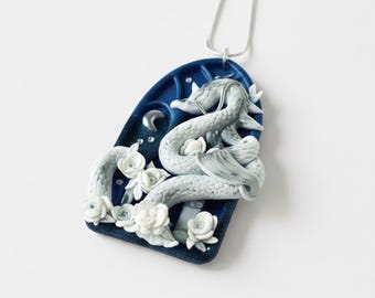 May include: A silver necklace with a pendant featuring a detailed, light gray dragon sculpture on a dark blue background. White flowers and a crescent moon accent the design. The pendant is approximately 2 inches long.