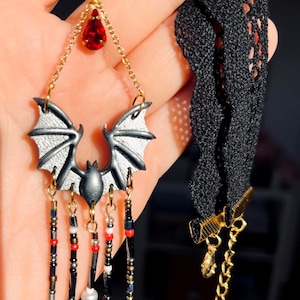 Choker hecho a mano con diseño de murciélago en tonos negro y gris, cuentas colgantes y lágrima roja, sobre fondo decorado con cuarzos, velas, piedras oscuras y detalles que imitan salpicaduras en rojo.