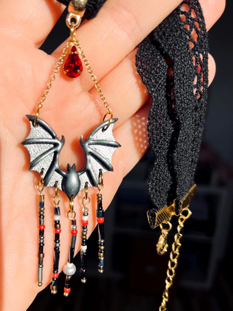 Choker hecho a mano con diseño de murciélago en tonos negro y gris, cuentas colgantes y lágrima roja, sobre fondo decorado con cuarzos, velas, piedras oscuras y detalles que imitan salpicaduras en rojo.