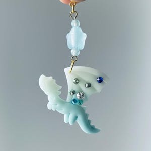 May include: A light blue dragon pendant with white wings, adorned with rhinestones and blue and silver beads. The dragon is suspended from a gold-colored chain with a light blue bead and a star-shaped bead.