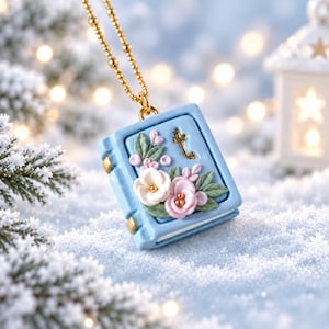 May include: A light blue book-shaped pendant necklace featuring a gold-tone chain. The cover is decorated with sculpted white and pink flowers, green leaves, and a small gold-tone letter t. The pendant has gold-tone metal accents on the spine and corners.
