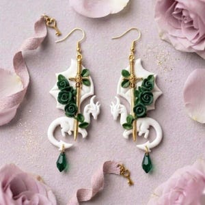 Puede incluir: Un par de pendientes de fantasía. Cada pendiente presenta un dragón blanco, una espada dorada y rosas verde oscuro. Una gema verde en forma de lágrima cuelga de la cola del dragón. Fondo rosa suave con pétalos.