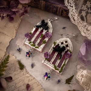 May include: A pair of stud earrings featuring a white gothic arch window frame with a translucent pink center. Each earring is adorned with a small black dragon figure, purple flowers, green vines, and a dangling iridescent star-shaped crystal charm.