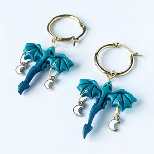 May include: A pair of gold hoop earrings with teal dragon charms and small white crescent moon charms. The dragon charms have detailed wings and tails. The earrings are displayed on a white background.